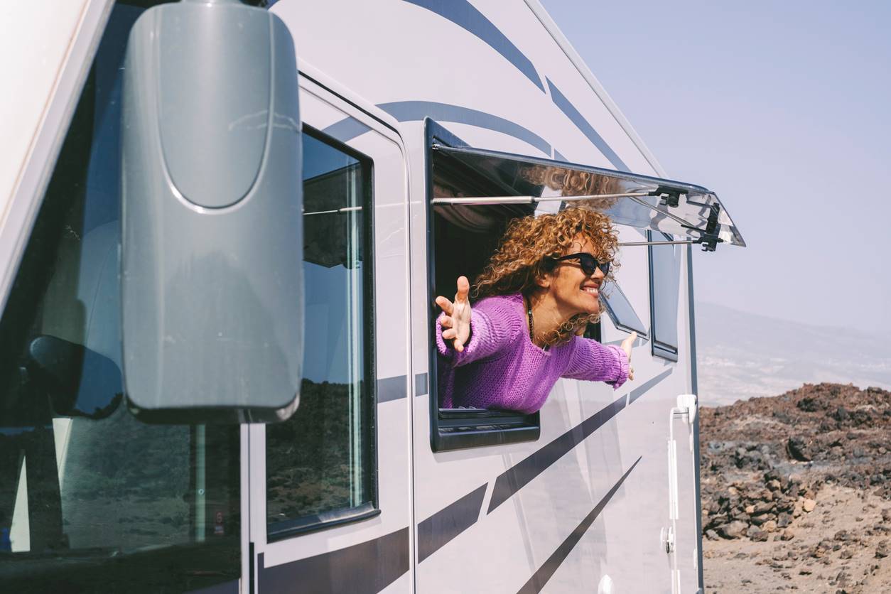 A smiling woman sticks her head and arms out of the window of an RV